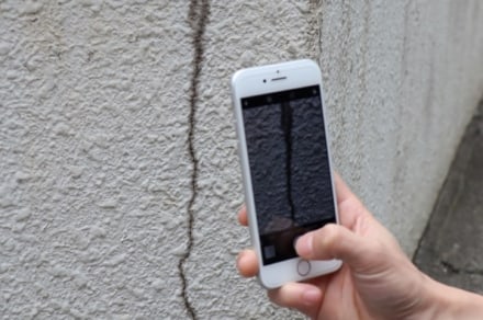 A photo of taking a picture of a crack on an exterior wall with their smartphone.